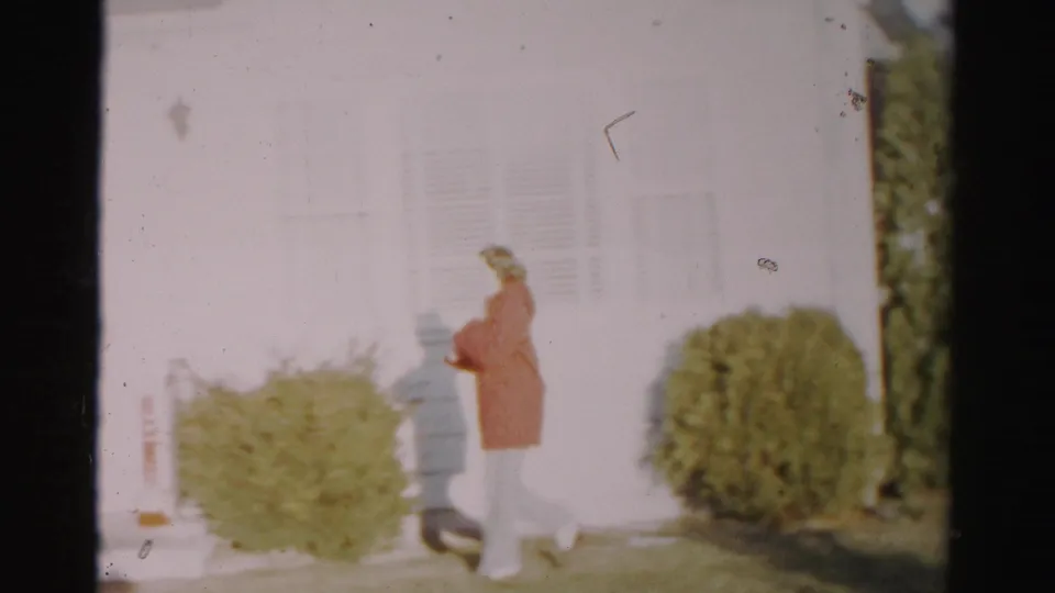 The woman nears the front door area of the Albany house, walking past low shrubs and white siding in soft, overexposed 8mm footage.