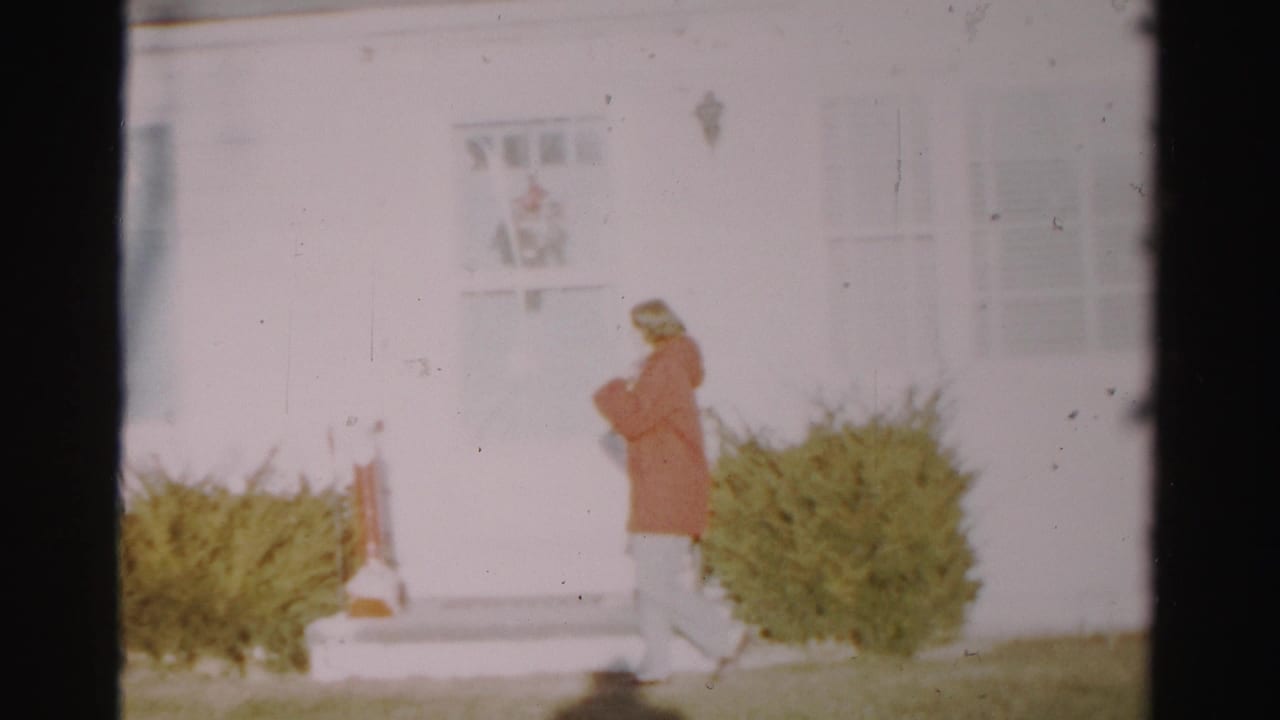 Near the end of the clip, the woman continues along the front of the white house, with shrubs, steps, and winter lawn visible.