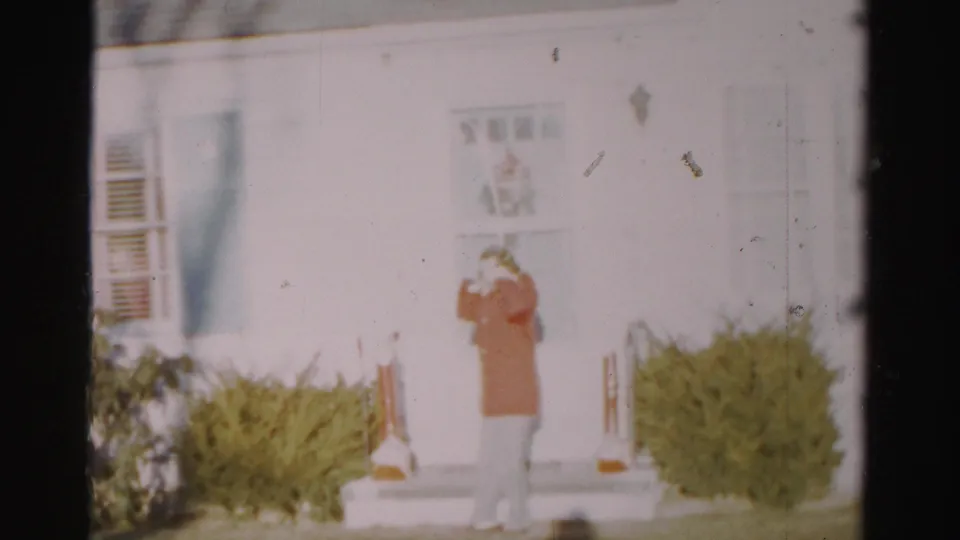 In Albany, New York home-movie footage, a woman in a red jacket stands outside a white house, lifting her hands near her hood.