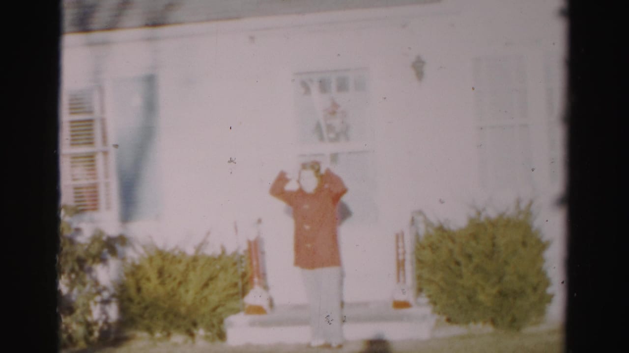 The woman stands centered on the steps of the Albany house, both hands raised as she adjusts a hood or head covering.