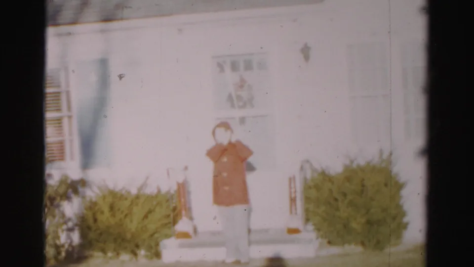 A soft 8mm frame shows the woman on the house steps in Albany, hand near her face after adjusting her head covering.