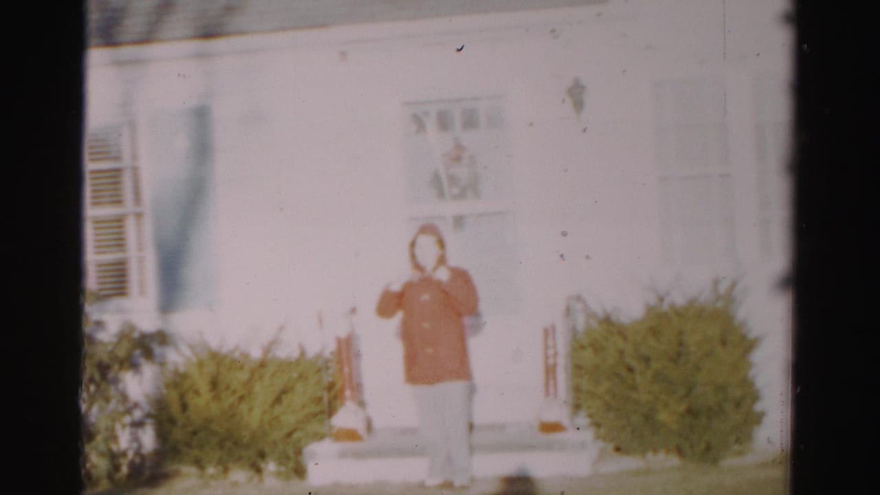 The woman stands outside the Albany home in a red jacket and light pants, framed by a white door, steps, porch light, and shrubs.