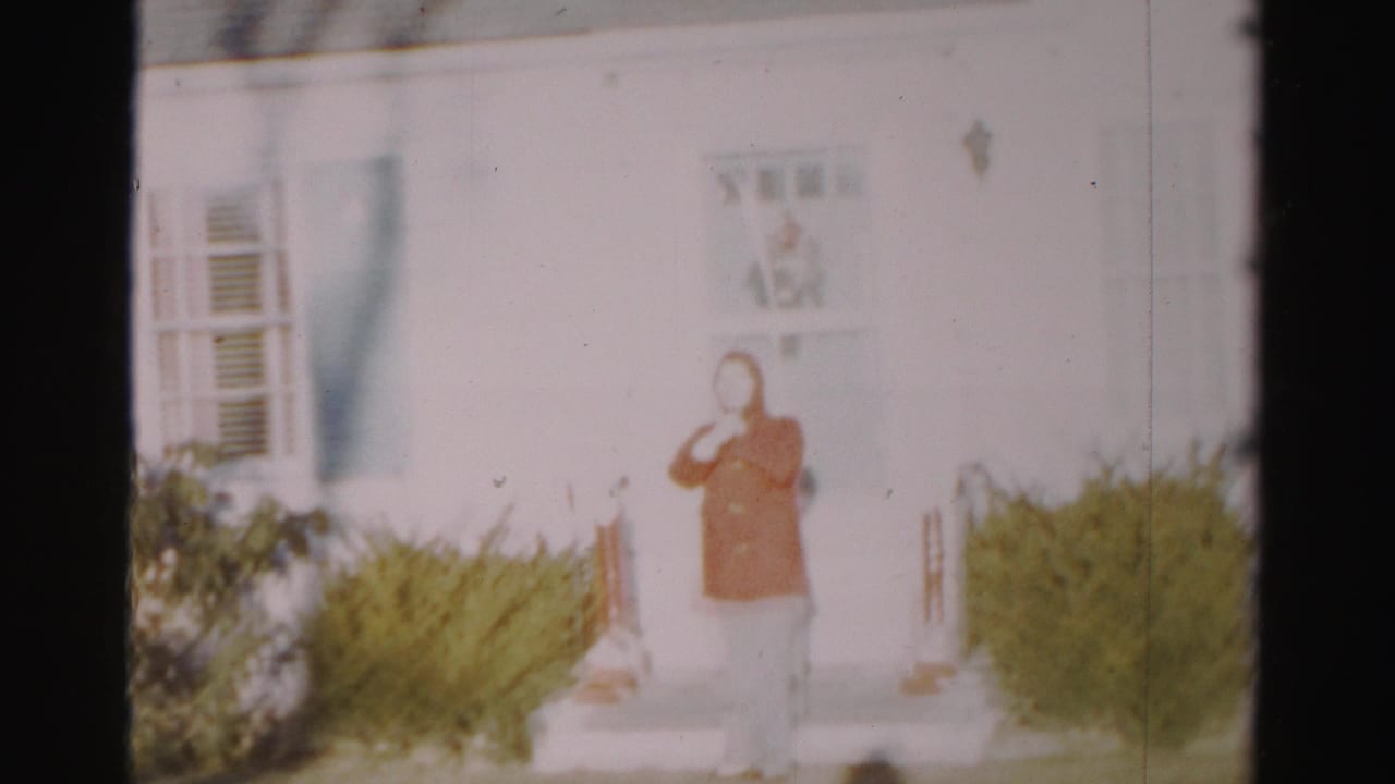 Person in a red coat stands on front steps of a white house with bushes nearby.