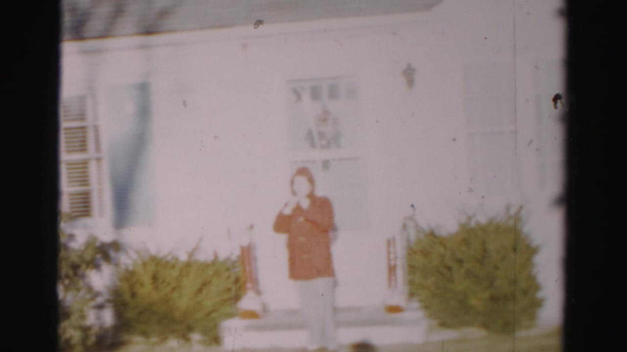 Person in red coat faces the camera from the steps of a white house.