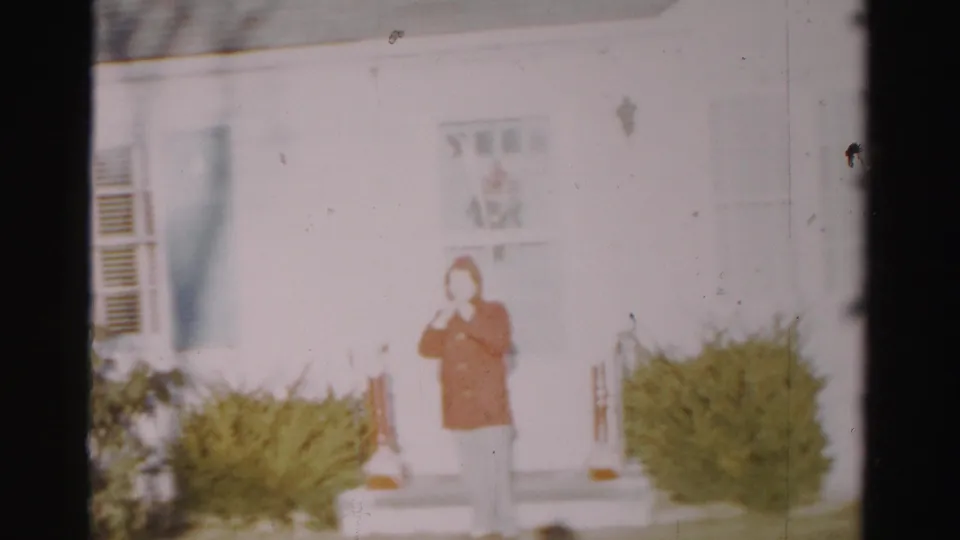 A person in a red coat and hat faces the camera on a front porch in Albany, New York, in 1950s 8mm footage.