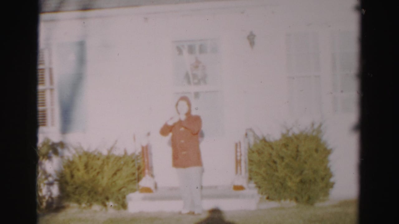 A soft-focus 1950s Albany frame shows a person in a red coat standing outside a white house entrance with bushes on both sides.