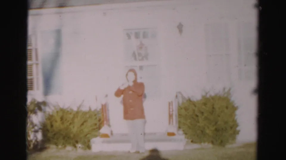 A soft-focus 1950s Albany frame shows a person in a red coat standing outside a white house entrance with bushes on both sides.