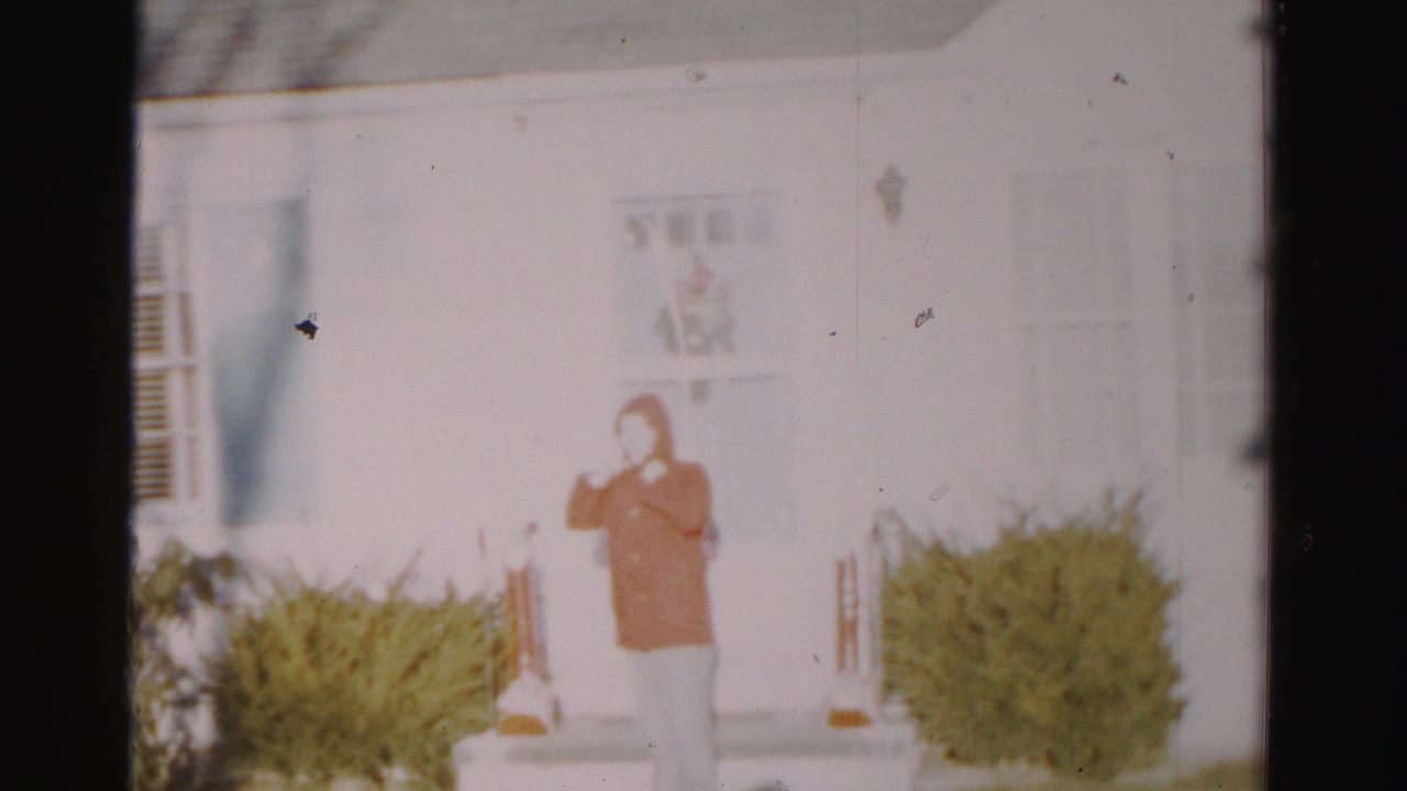 Adult in an orange hooded raincoat stands on a porch in front of a white house.