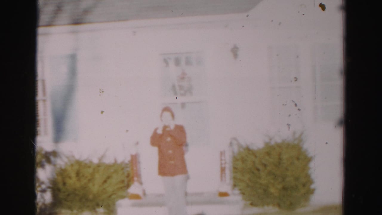 Adult in orange outerwear stands centered outside a white house with shrubs nearby.