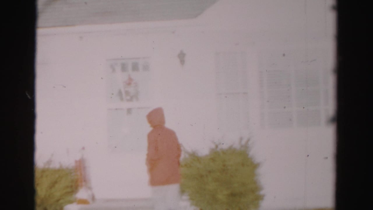 The hooded adult turns away toward the entrance of the Albany house, with snow and shrubs visible in soft-focus 8mm footage.