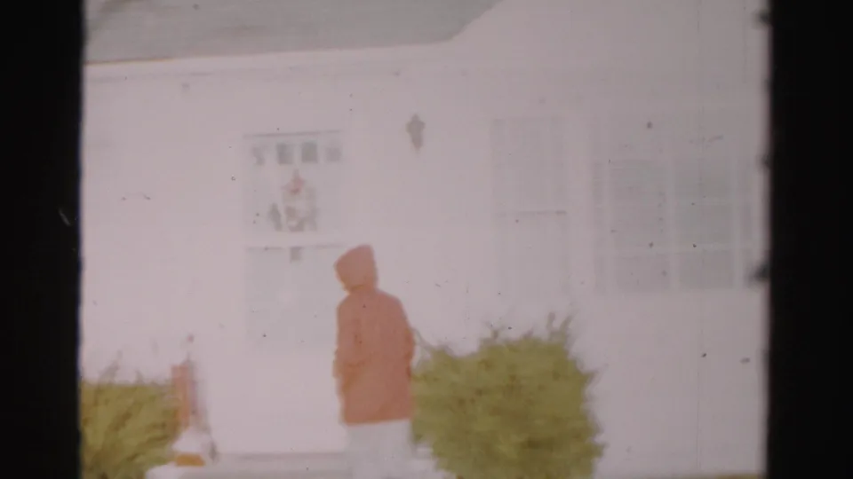 The hooded adult turns away toward the entrance of the Albany house, with snow and shrubs visible in soft-focus 8mm footage.