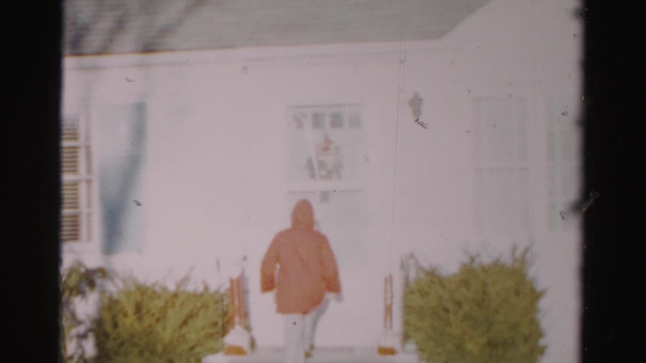 The adult in an orange raincoat walks toward the front door of the white Albany residence as the home movie frame drifts slightly.