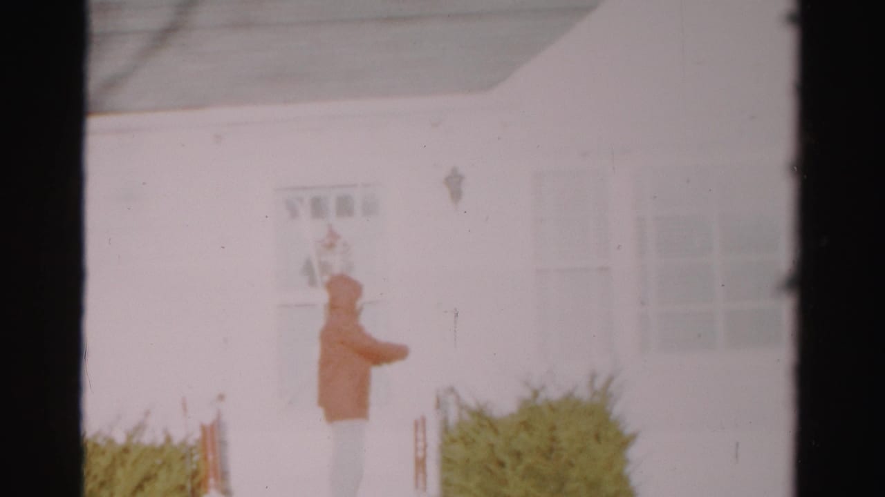 Hooded person reaches toward the door area of a white house.