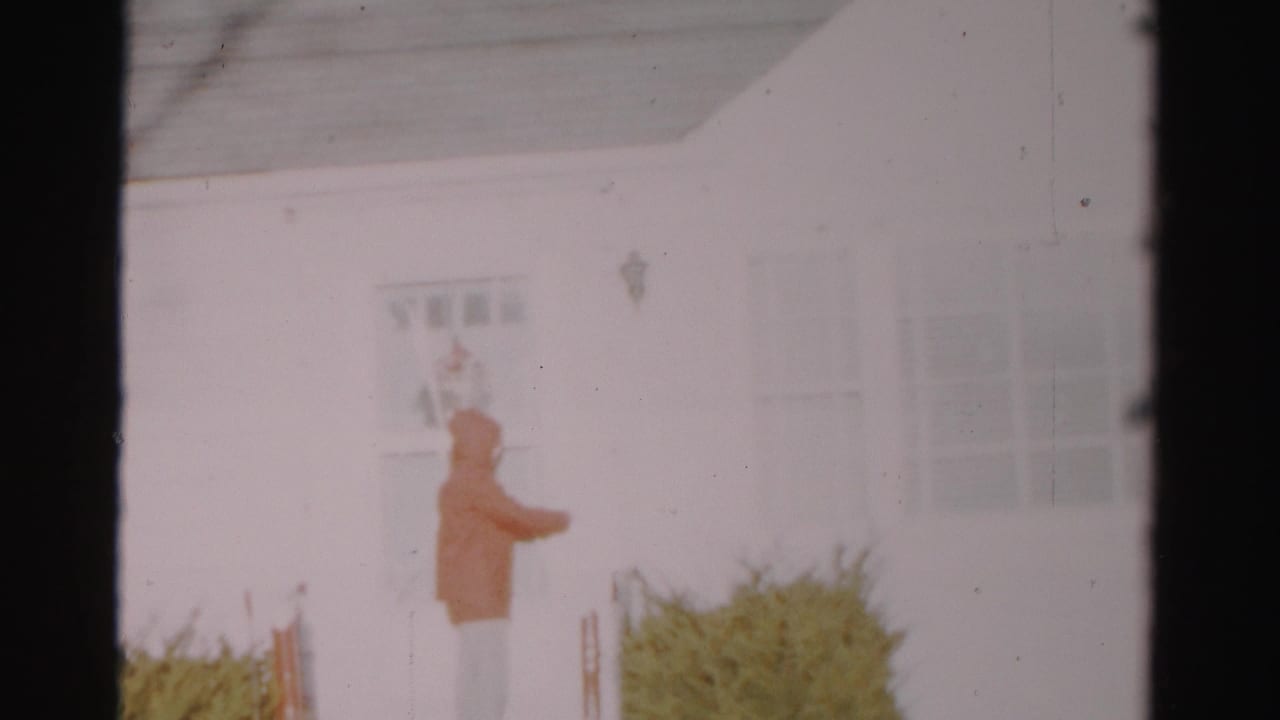 The 1956 Albany home-movie frame shows a hooded visitor paused beside the door and low shrubs of a white house.