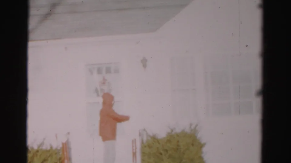 The 1956 Albany home-movie frame shows a hooded visitor paused beside the door and low shrubs of a white house.