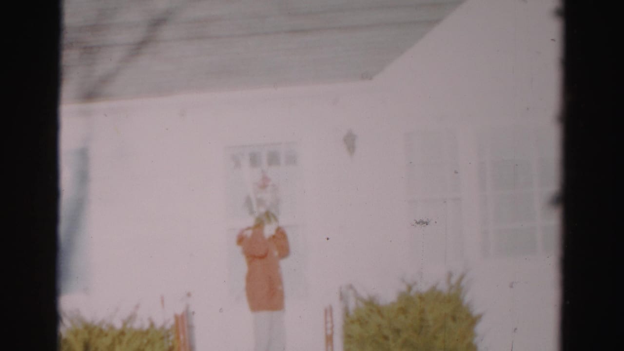 A hooded person stands close to the door of a white suburban house, with windows and shrubs visible in the soft 8mm frame.
