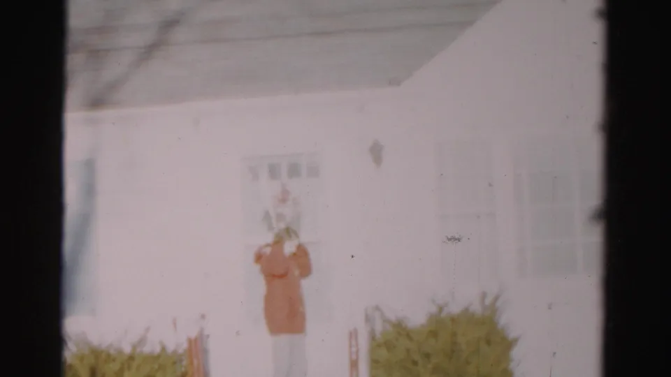 A hooded person stands close to the door of a white suburban house, with windows and shrubs visible in the soft 8mm frame.