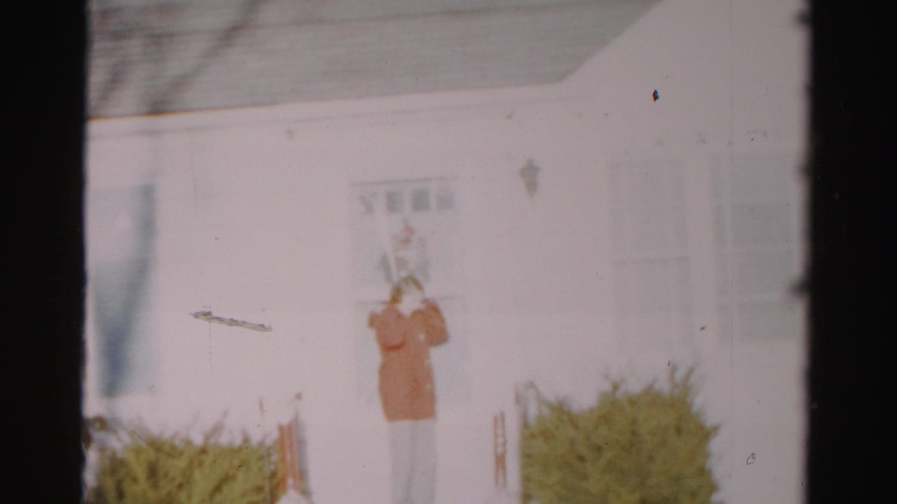 A woman in a red coat stands on the porch area of a white Albany-area house in archival 8mm footage from the 1950s.
