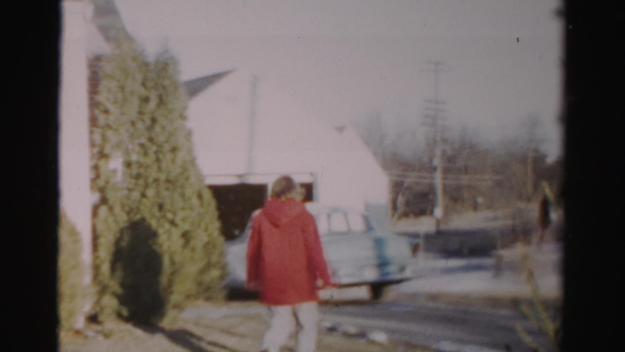In the Albany home-movie scene, the woman walks from the house area toward a pale car near a garage and utility poles.
