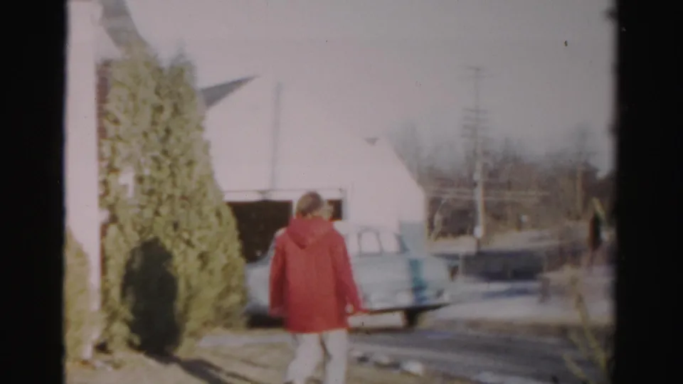 In the Albany home-movie scene, the woman walks from the house area toward a pale car near a garage and utility poles.