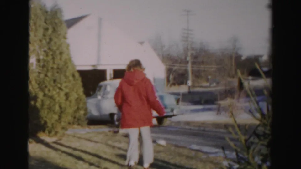 The woman continues across the yard or driveway area, with a mid-century car, garage, utility poles, shrubs, and winter-bare trees visible.