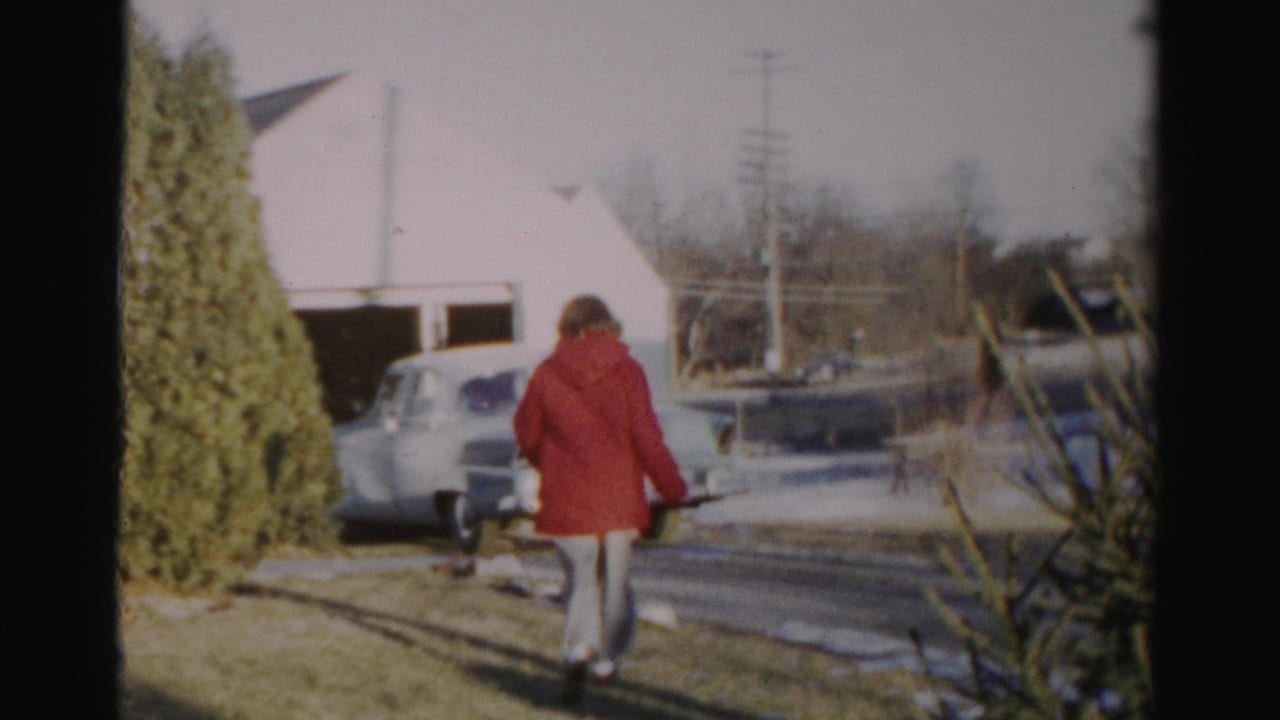 In 1950s Albany home-movie footage, a person in a red coat walks over a patchy snowy lawn toward a pale vintage car near a house or garage.