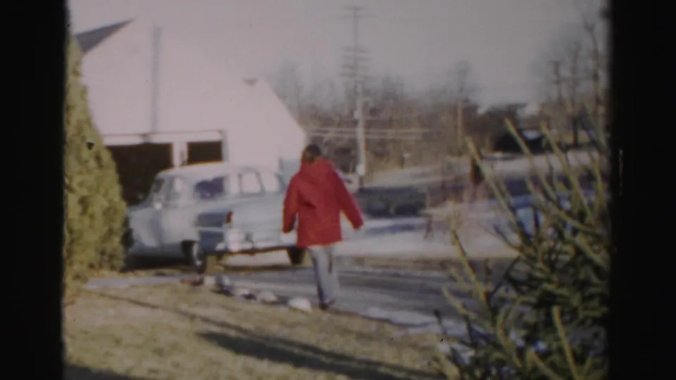 A red-coated person moves closer to a pale mid-century car in a snowy suburban yard, with shrubs, bare branches, and utility poles behind.