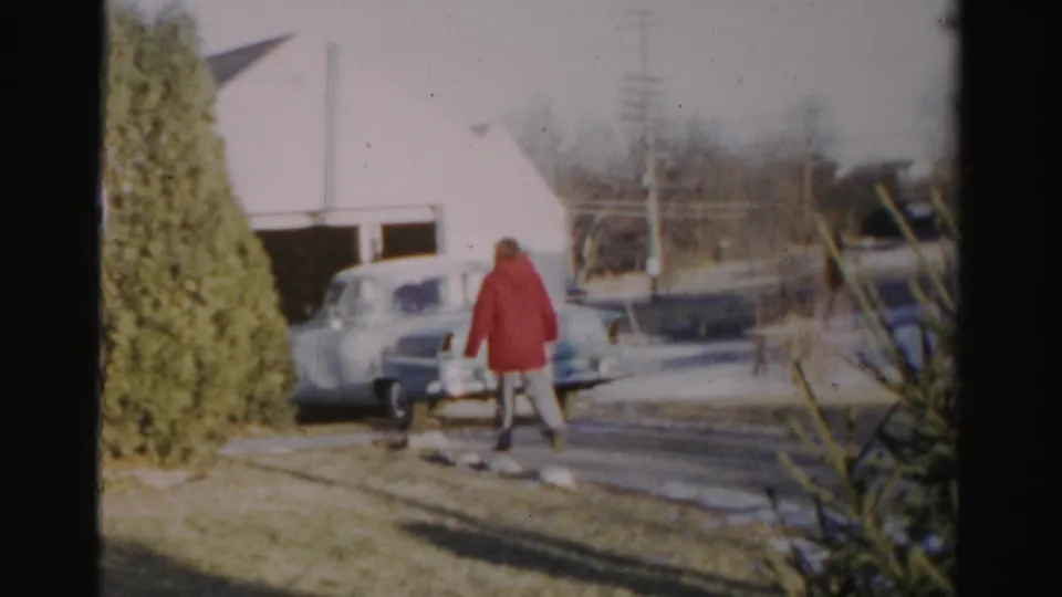 The person in the red coat reaches the area near the rear of the pale vintage car, with a winter lawn and residential structures visible around them.