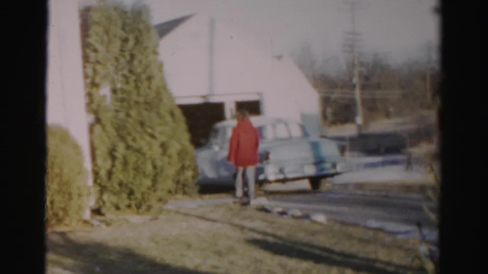 A person in a red coat stands beside or just behind the pale vintage car in a patchy snowy Albany residential yard, filmed in soft 8mm color.