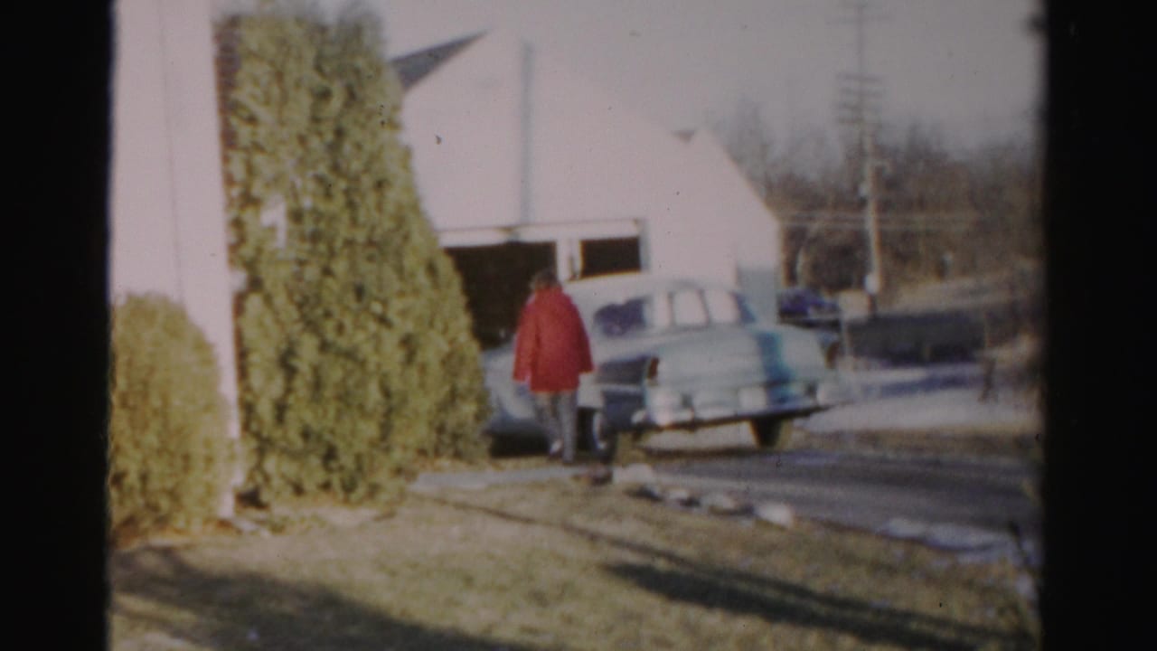 In 1956 Albany home-movie footage, an elderly woman in a red jacket approaches a pale blue car beside a suburban garage.