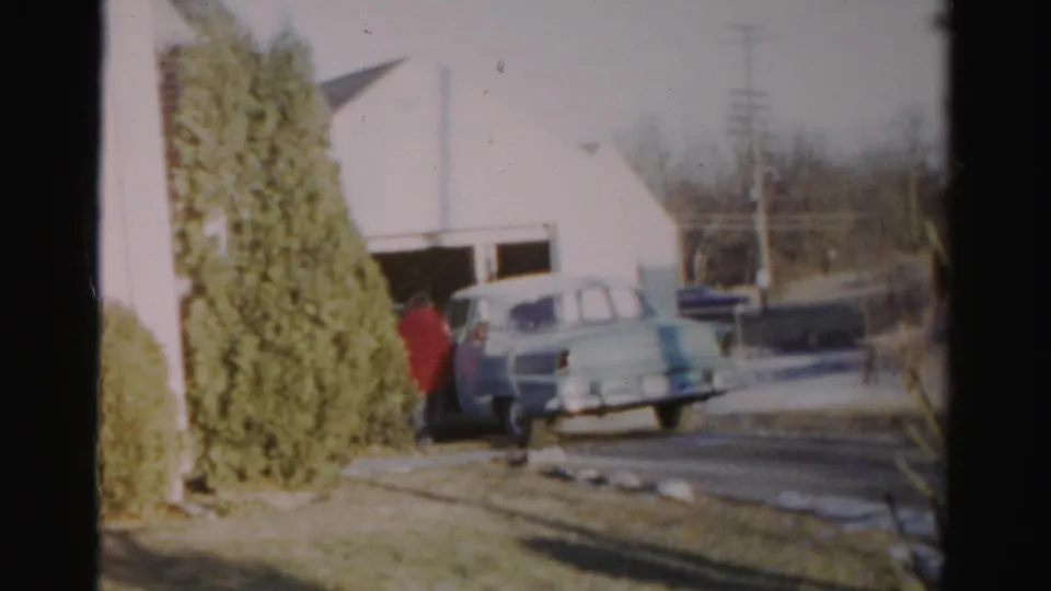 At the end of the clip, the woman remains beside the pale blue car in the 1956 Albany residential scene.