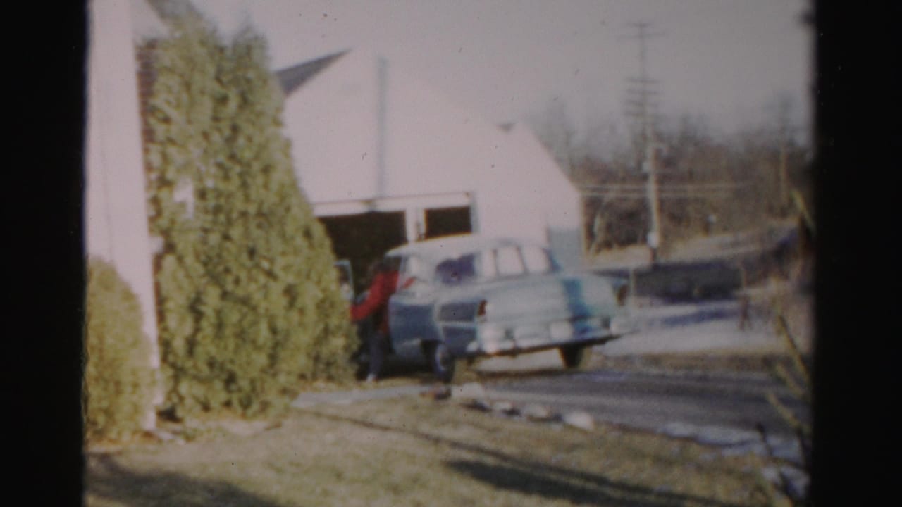 A person in red stands by a light blue car near a garage in Albany-area 1950s home-movie footage.