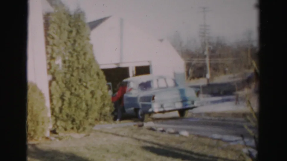 A person in red stands by a light blue car near a garage in Albany-area 1950s home-movie footage.