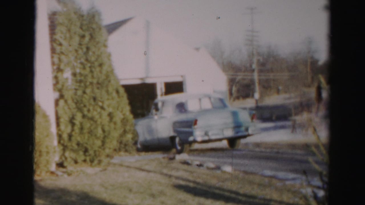 1956 Albany home-movie frame showing a light blue mid-century sedan reversing near a white garage and evergreen shrub.