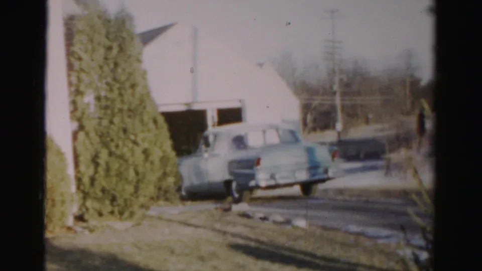 1956 Albany home-movie frame showing a light blue mid-century sedan reversing near a white garage and evergreen shrub.