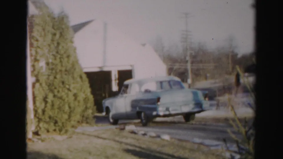 1956 Albany home-movie frame showing the sedan near the driveway entrance, with lawn stones and garage visible.
