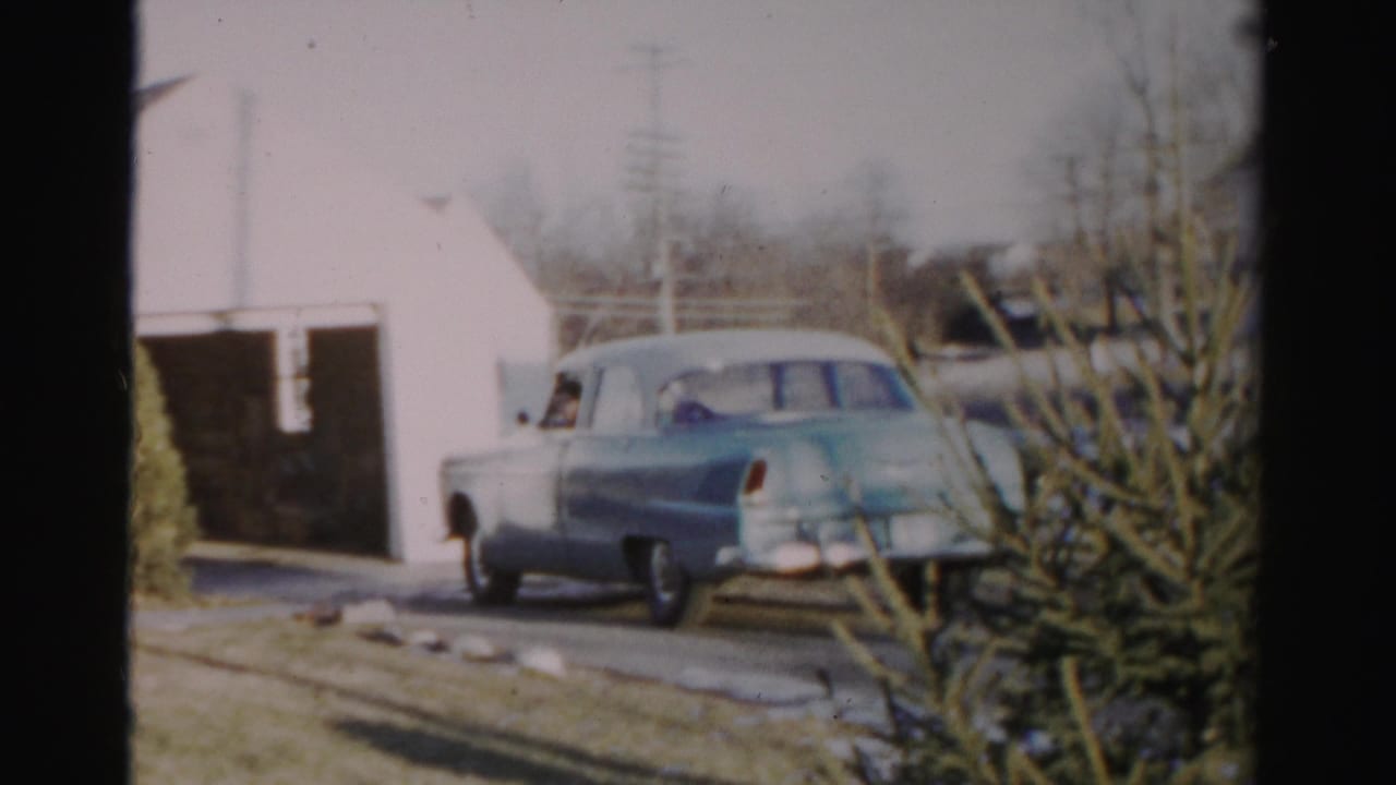 Light blue 1950s sedan backing from a driveway beside a white garage and evergreen shrub.