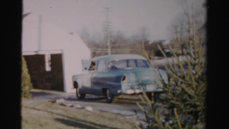 In Albany, New York home-movie footage, a light blue mid-century sedan reverses from a residential driveway near a white garage.