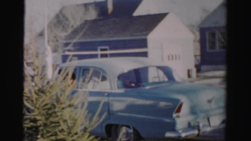 The final frame shows the rear of the mid-century sedan near the driveway, with residential buildings and an evergreen shrub in the foreground.