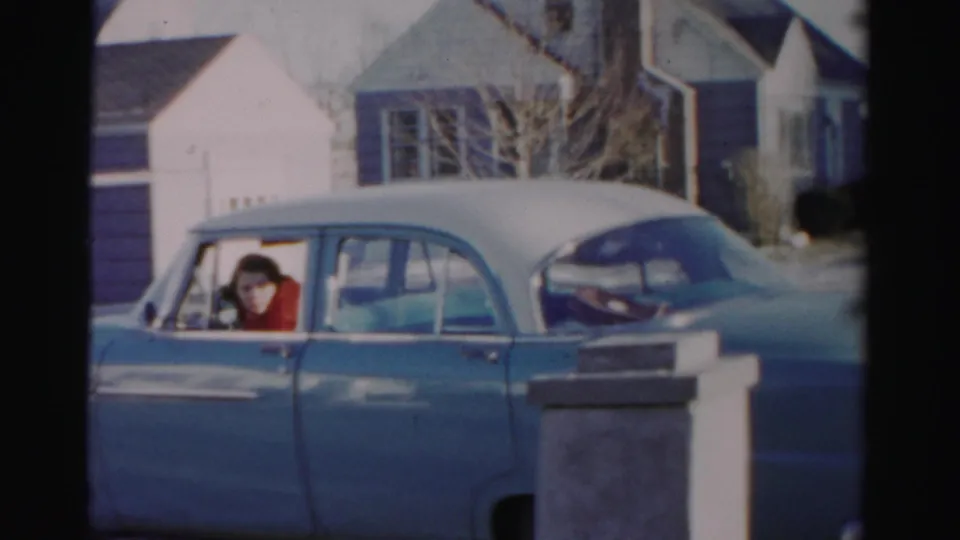 A light blue sedan with a white roof passes a house exterior in Albany, with the woman driver visible through the side window.