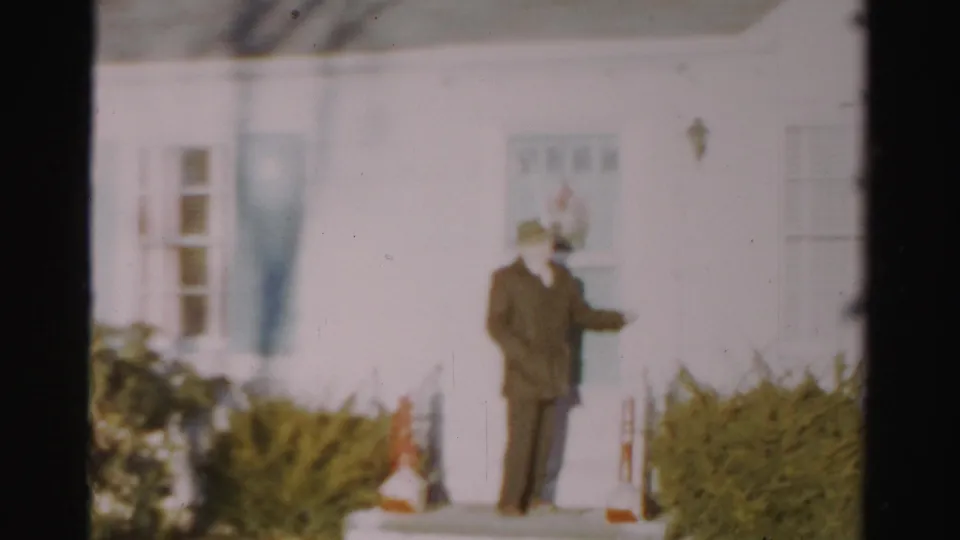 1956 Albany frame showing a man in a dark suit and hat near a white house entrance with a wreath and winter shrubs.