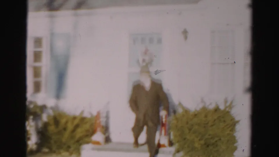 A man in a dark suit and hat walks down from a white house entrance in a soft 1950s Albany home-movie frame.