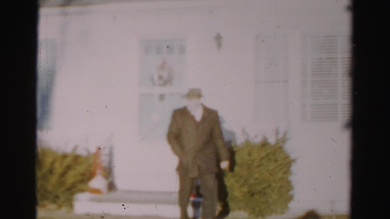 In 1950s Albany home-movie footage, a man in a fedora and dark coat stands or walks near white siding and clipped shrubs.