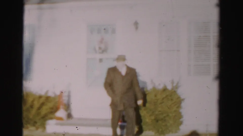 In 1950s Albany home-movie footage, a man in a fedora and dark coat stands or walks near white siding and clipped shrubs.