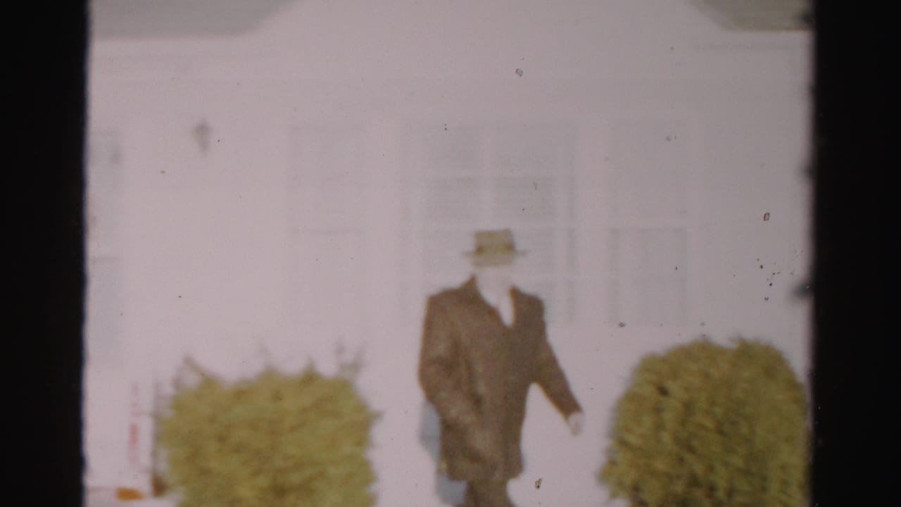 The man in a fedora and dark coat walks across a residential yard in Albany, with pale siding and shrubs behind him.