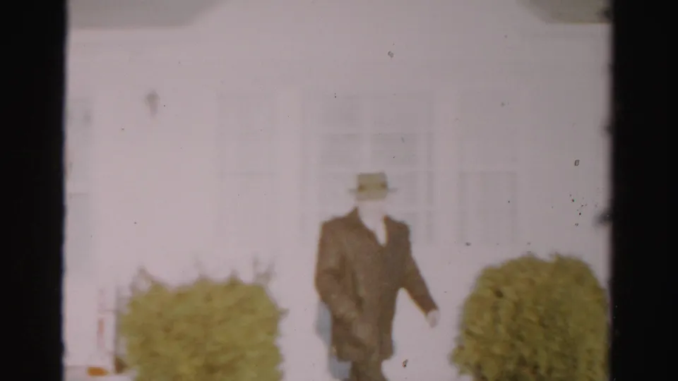 The man in a fedora and dark coat walks across a residential yard in Albany, with pale siding and shrubs behind him.