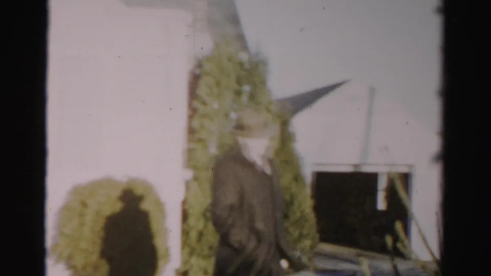 A close, soft 8mm frame shows the fedora-wearing man moving past shrubbery beside a residential exterior in Albany.