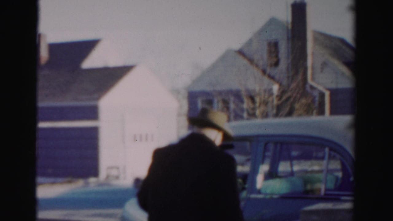 Man in a dark coat stands beside a blue car on a snowy residential street.