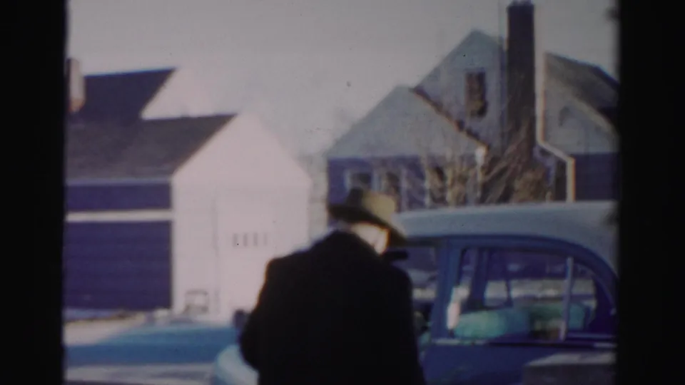 A man in a fedora and overcoat stands by a blue mid-century car on a snowy Albany residential street.