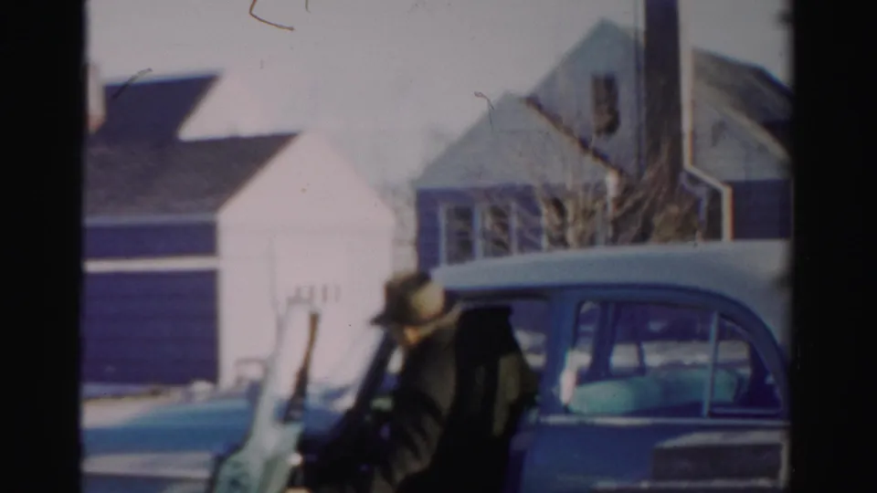 The man leans toward the blue car as if opening or entering it, with snow and houses visible behind him.